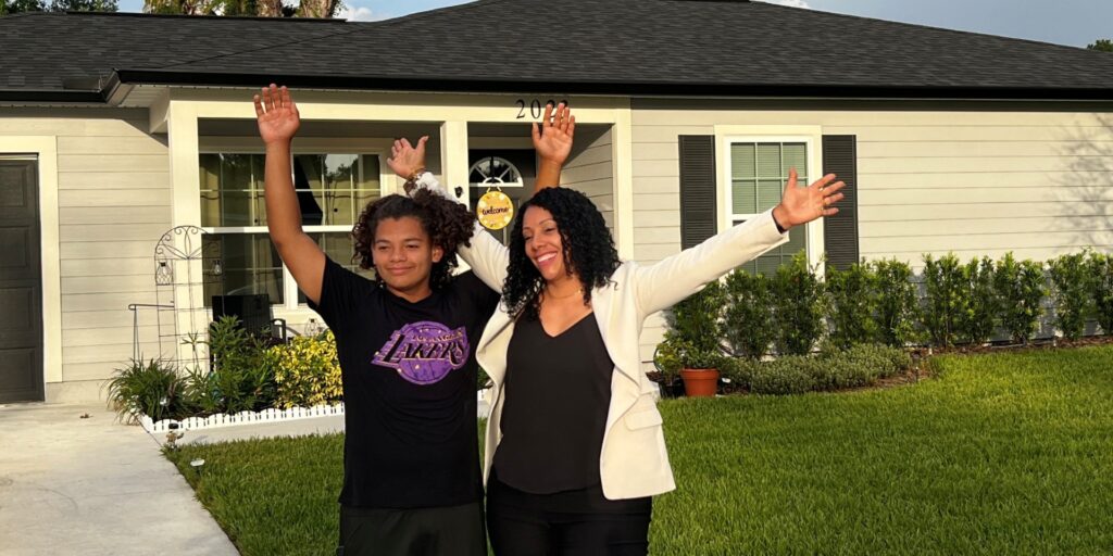 A mother and son stand in frotn of their home with their arms raised in celebration at sunset